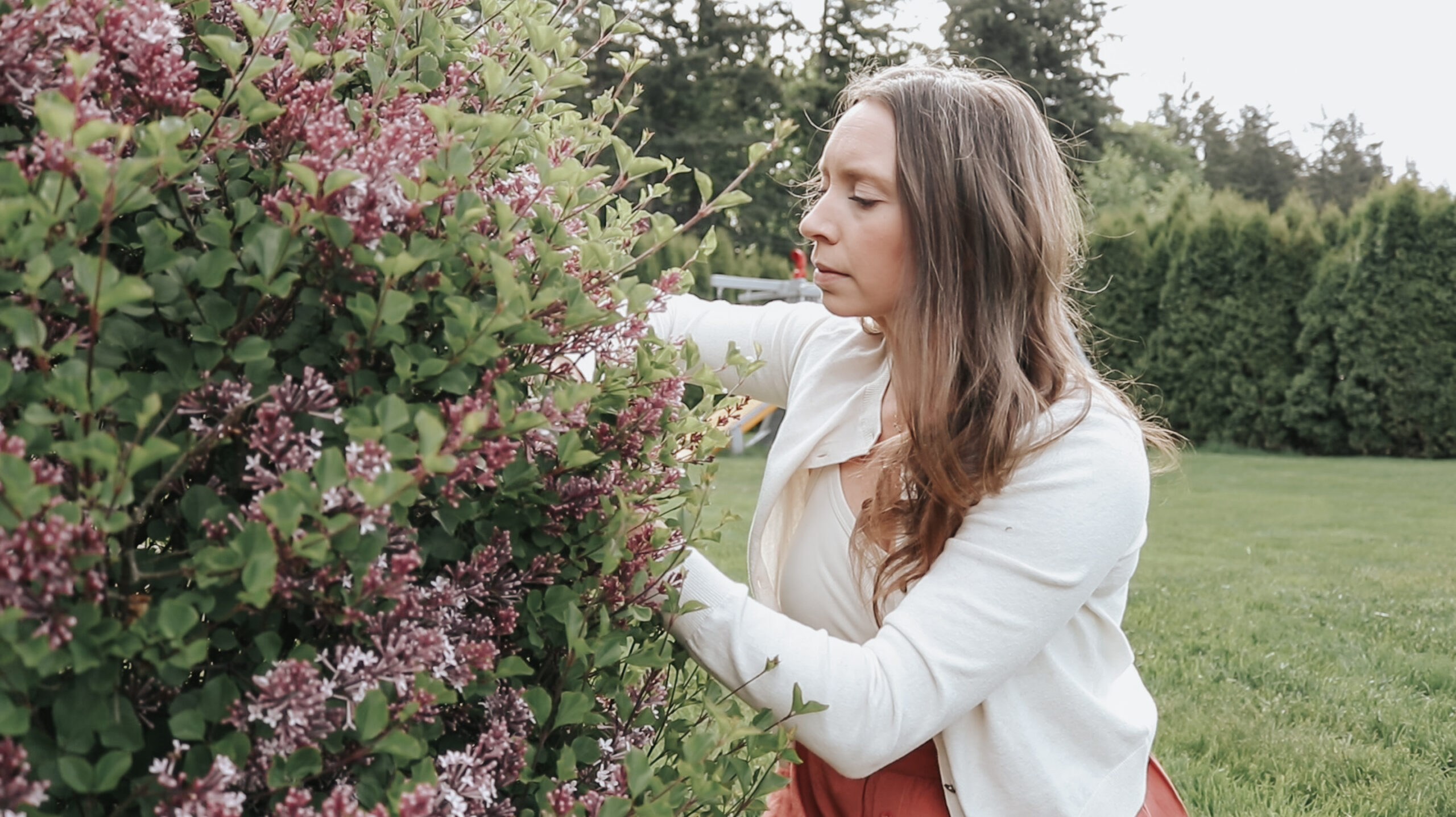 Christian homemaker trims flowers from a tree outside. She is wearing a white cardigan and red skirt.